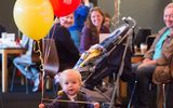 A young toddler is walking towards the camera. She is holding a bunch of red and yellow balloons. Three adults are in the background smiling at her.