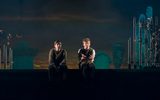 A theatrical production image. Two young men in scruffy 50s style clothing sit on the edge of the stage, which is lit dramatically and features a backdrop showing a sunset.