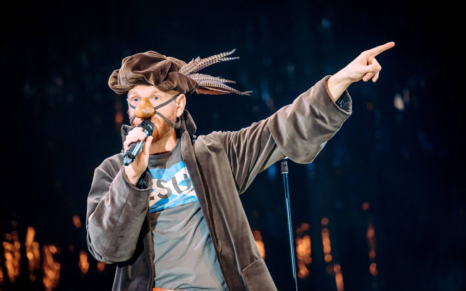 A theatrical production. A man wearing a brown highlander-style cap and a fake nose speaks into a microphone and points to the right.