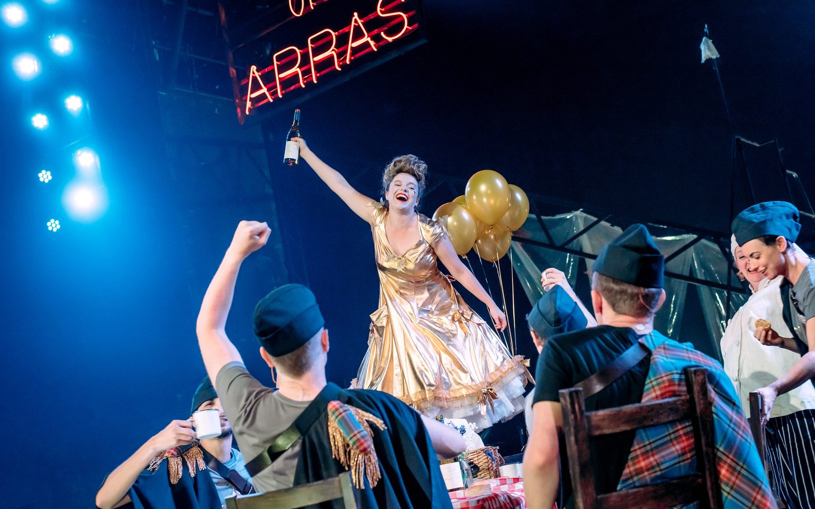 A theatrical production. A woman is surrounded by six actors sitting at a table. The woman is wearing a gold dress, standing on the table and holding a bottle of wine in the air.