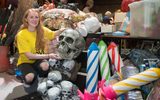 A sale of props and other theatrical items. A girl is holding a large prop skull and smiling at the camera. There are large, colourful candles next to her.