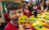 A classroom. A group of children are sitting at a table. One child is holding up a yellow emoji sticker to the camera and sticking his tongue out.