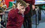 A classroom. A young boy in a dark red school jumper is making a funny face and holding his hands up to his head.