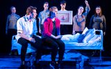 A theatrical production. A young woman lies in a hospital bed. A second woman is perched on the side of the bed and a young man is standing talking to them holding a clip board. They are surrounded by a group of people all wearing striped tops. One of the group is holding a painting of a yacht and another is holding a up an IV drip.