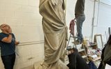 A statue of a Greek Goddess in a workshop. A man is standing on a ladder beside the statue measuring it. Other people are standing beside looking up at the figure.