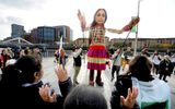 A large wicker puppet of a young girl stands looking down at a long line of school children who are walking past her, smiling and waving.