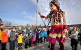 A large wicker puppet of a young girl stands looking down at a long line of school children who are walking past her, smiling and waving.