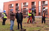 Two men in warm jackets and hats are standing next to each other. Thy are looking at something off camera. In the background are other people warm clothing, high visibility vests and hard hats. One man in the background is being filmed by a camera crew