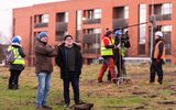 Two men in warm jackets and hats are standing next to each other. They are looking at something off camera. In the background are other people warm clothing, high visibility vests and hard hats. One man in the background is being filmed by a camera crew