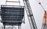 a large crane is pictured lifting a large metal structure into the air. The sky is blue with clouds.