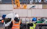 a large crane is pictured lifting a large metal structure into the air. The sky is blue with clouds. Two people in high visibility vests and hard hats are looking at the construction site.