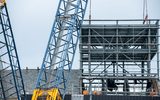 a large crane is pictured lifting a large metal structure into the air. The sky is blue with clouds.
