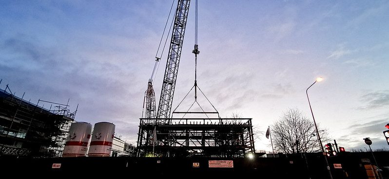 a large crane is pictured lifting a large metal structure into the air. The sky is blue with clouds.