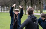 A young person with glasses, dressed in a jacket and cap, smiles and gives a thumbs up to the camera.