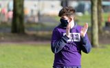 A young person wearing a mask and a purple Citizens Theatre t-shirt claps their hands while looking to the left. They are in a park.