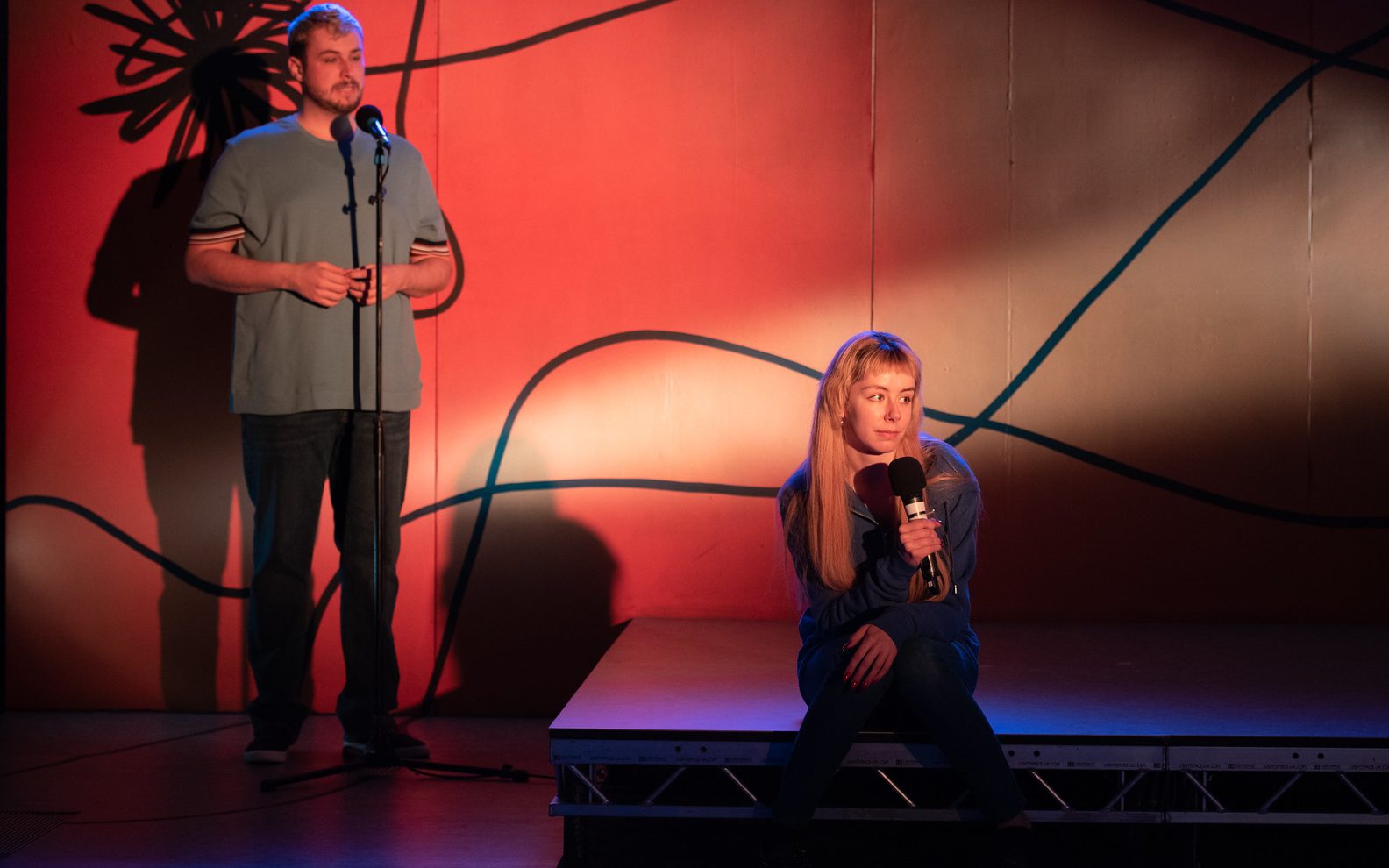 A stage show. A man is standing in front of a microphone. A woman is sitting on the floor in front of him holding a microphone.