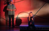A stage show. A man is standing in front of a microphone. A woman is sitting on the floor in front of him holding a microphone.