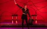 A stage show. A woman is standing on stage. She is wearing a school uniform. She is waving a vodka bottle in the air and holding a backpack in the other hand.
