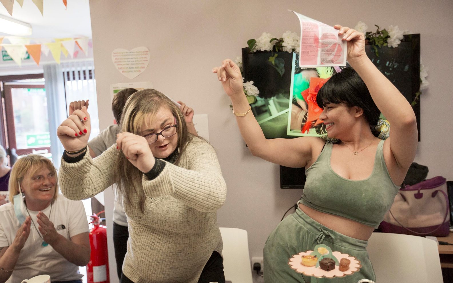 Two women smiling and dancing at an All We Can Do sharing session. Other women can be seen clapping in the background.]