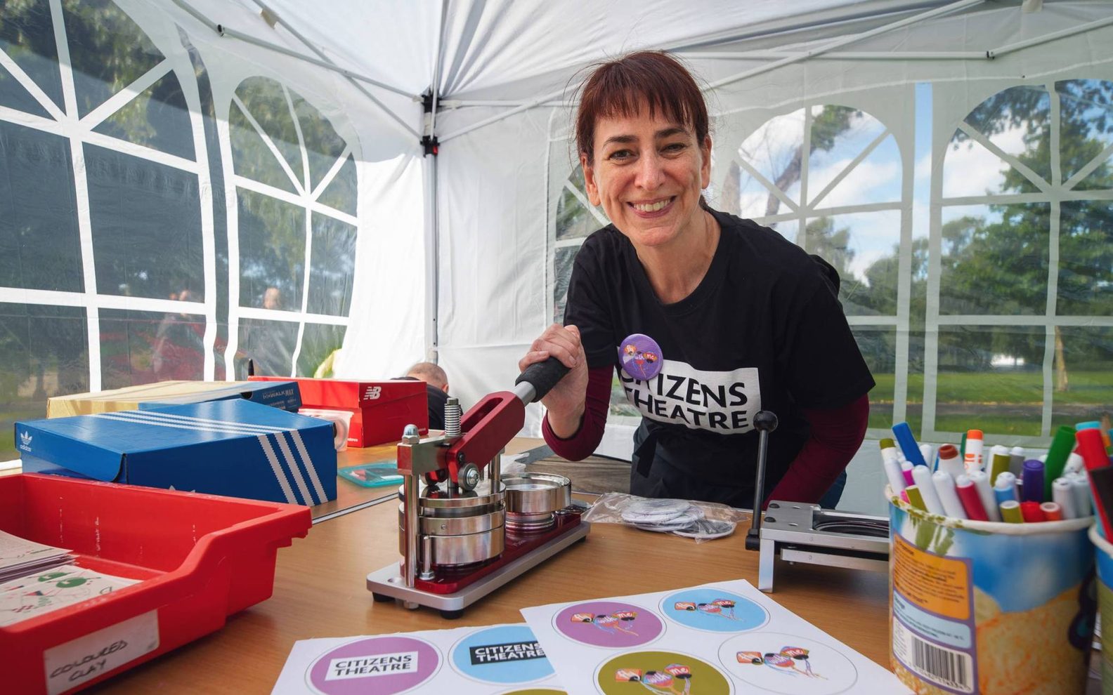 Citizens Theatre staff member Elly Goodman smiles at the camera. She is in a tent and using a button badge making machine.
