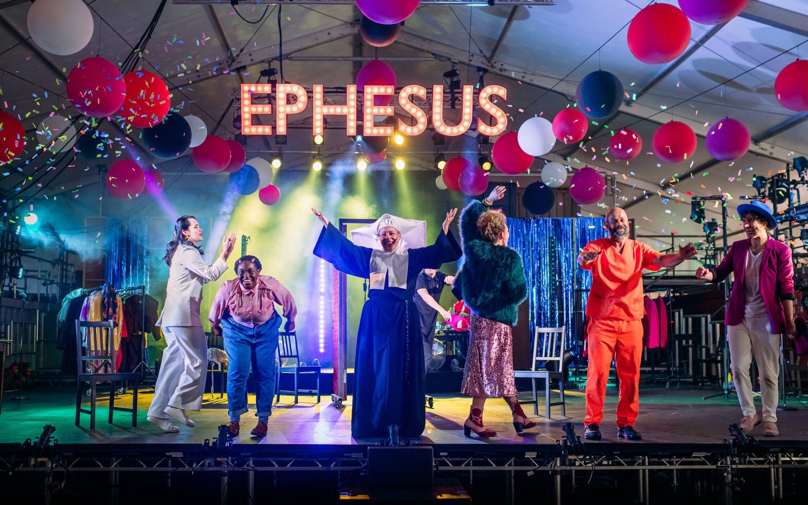 A highly theatrical production shot. Six figures are on a stage, dressed in colourful clothing. They are smiling and gesturing as in celebration, and confetti is falling. The word 