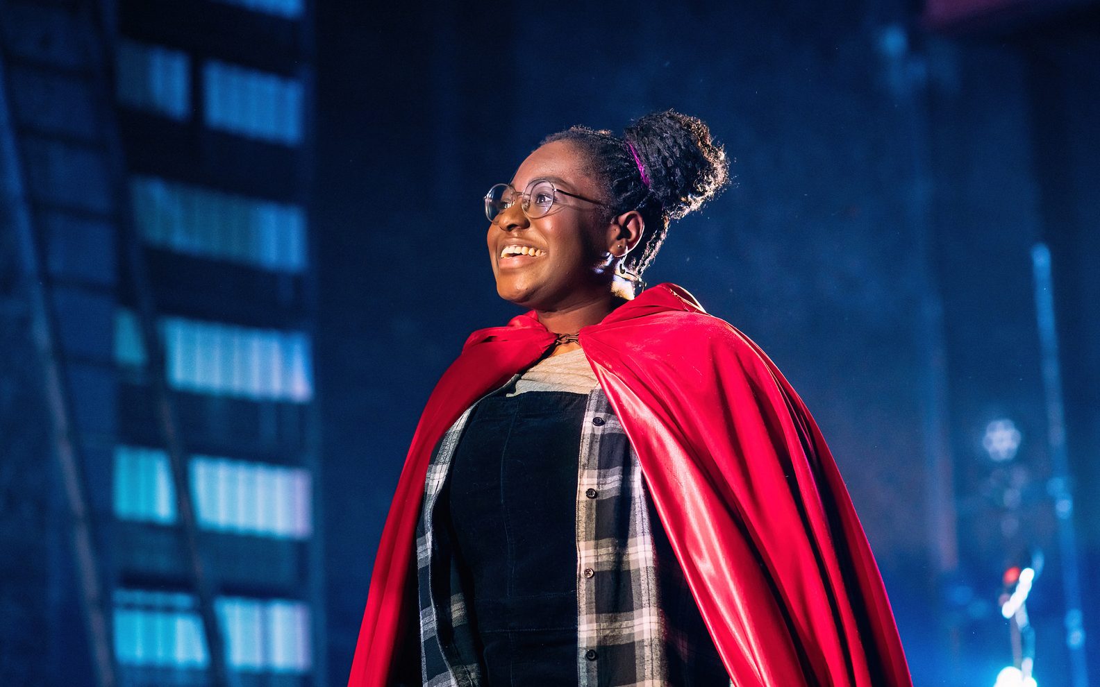 A girl with glasses stands on stage, smiling. She is wearing a bright red cape over her modern clothes.