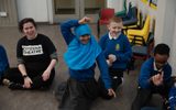 Primary pupils sitting on the floor. They are smiling. A woman in a Citizens Theatre t-shirt is sitting next to them.