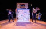 A stage show. Two actors standing next to a large map. They have backpacks on and are wearing binoculars. They both have one hand on their hip and the other held up to their forehead as if looking out to the distance.