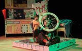 A stage show, there are two actors sitting in a large wooden drawer. One is holding up a wheel and looks like she is steering. The other is sitting behind holding up a flag.