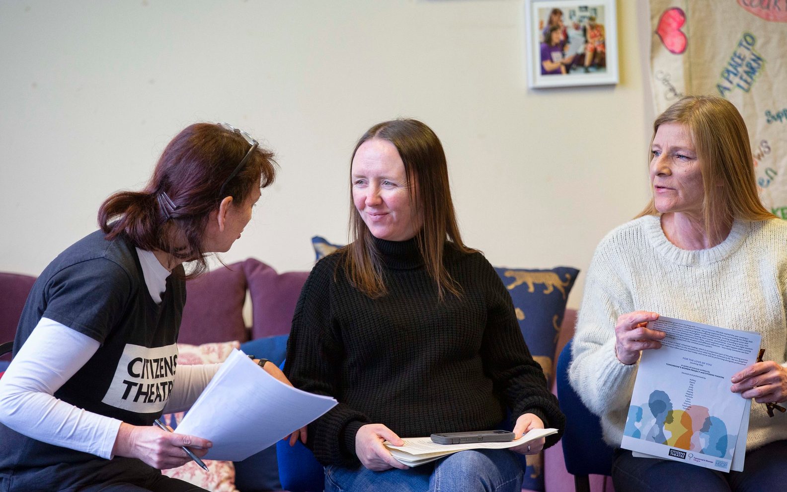 A woman wearing a Citizens Theatre t-shirt talks to a young woman. Both are sitting down and holding scripts.