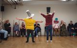 Two adults stand with their backs to the camera, gesturing broadly with their arms. They are wearing large hats made out of cardboard. Several other people sit in a row in front of them applauding.