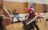Two adults in large colourful hats pose while sitting on a black box. Others can be seen applauding. One has a large keyboard prop.