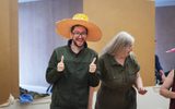 Two adults standing in a room. One is wearing a large paper hat and smiling to the camera while giving a thumbs up.