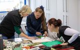 Three women are around a table. They are surrounded by art materials. They are looking at something on the table.