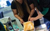 Two women are standing together. They are lifting a piece of paper to reveal a printed image.