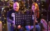 A man and a woman are sitting on a sofa. They are wearing Citizens Theatre tshirts. The woman is holding a guitar. They appear to be singing.