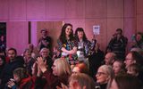 A busy audience clapping. Two performers are walking through the audience wearing lots of festive sparkles.