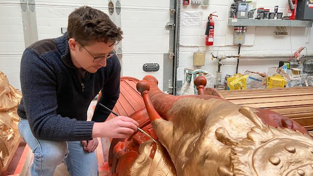a man in a navy jumper and jeans is crouched beside a statue. He has a long thin paintbrush and is painting the statue with gold paint