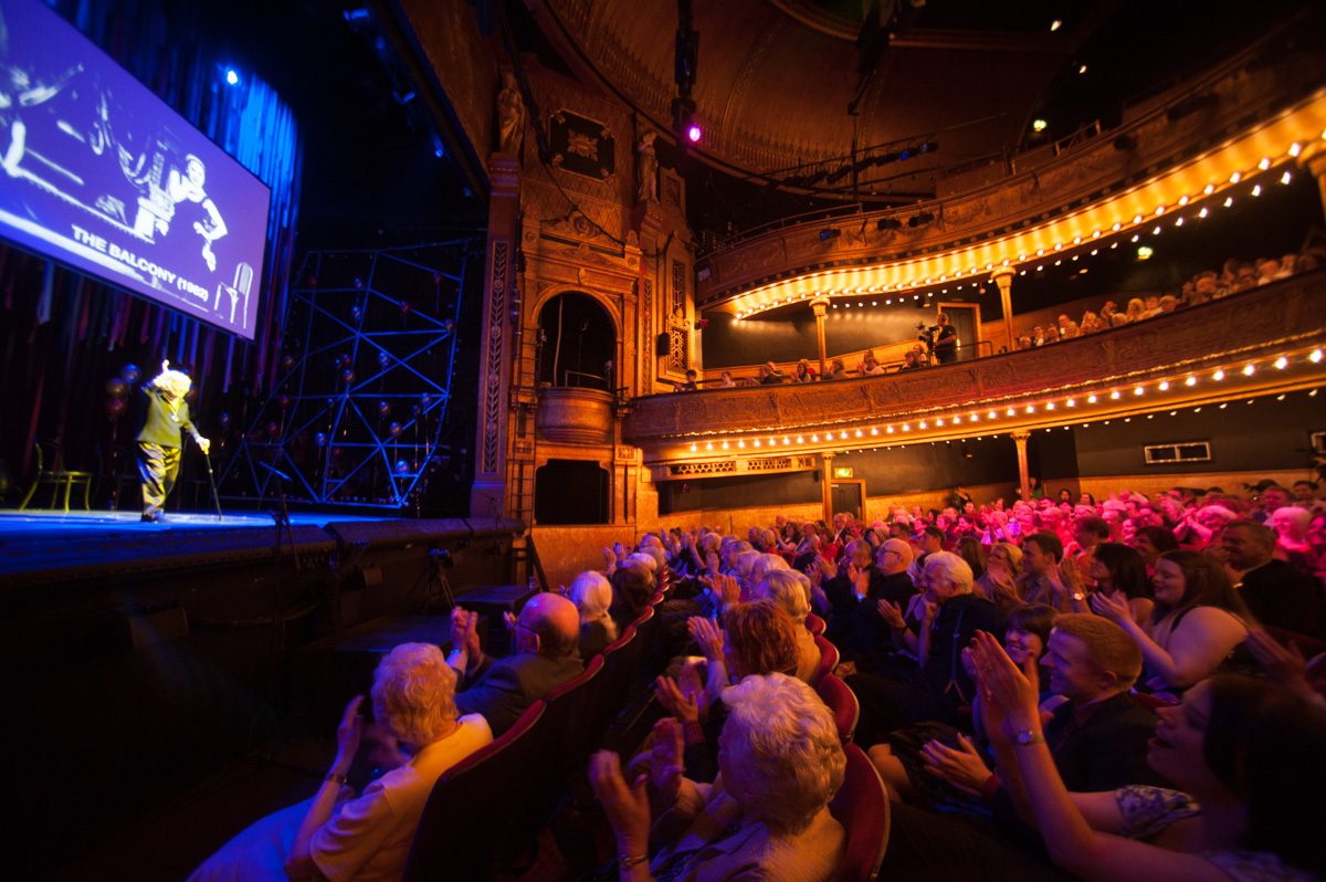Side view of the auditorium of the Citizens Theatre during a performance. A person is on stage and a full audience is clapping.