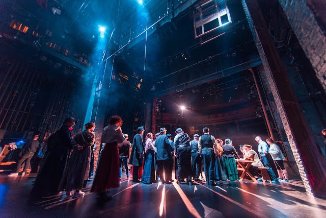 A large cast of people dressed in early 20th century costumes, taken from the back of the stage facing the audience.