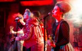 A highly theatrical production image of a show. Everybody is wearing punk style clothing. They are on a stage with graffiti style surroundings. A band are singing into microphones and playing guitars.