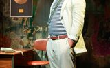 A highly theatrical production image of a show. Everybody is wearing punk style clothing. They are on a stage with graffiti style surroundings. A man in a white suite and stripe tshirt is standing with his hands in his pockes.