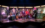 A highly theatrical production image of a show. Everybody is wearing punk style clothing. They are on a stage with graffiti style surroundings. A group of yound people are all standing on the stage looking at one person at the end of a line.