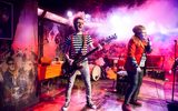 A highly theatrical production image of a show. Everybody is wearing punk style clothing. They are on a stage with graffiti style surroundings. A band are playing guitars and singing into microphones.