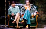 A highly theatrical production image of a show. Everybody is wearing punk style clothing. They are on a stage with graffiti style surroundings. A boy in a hooded anorak is sitting on a couch with a woman.