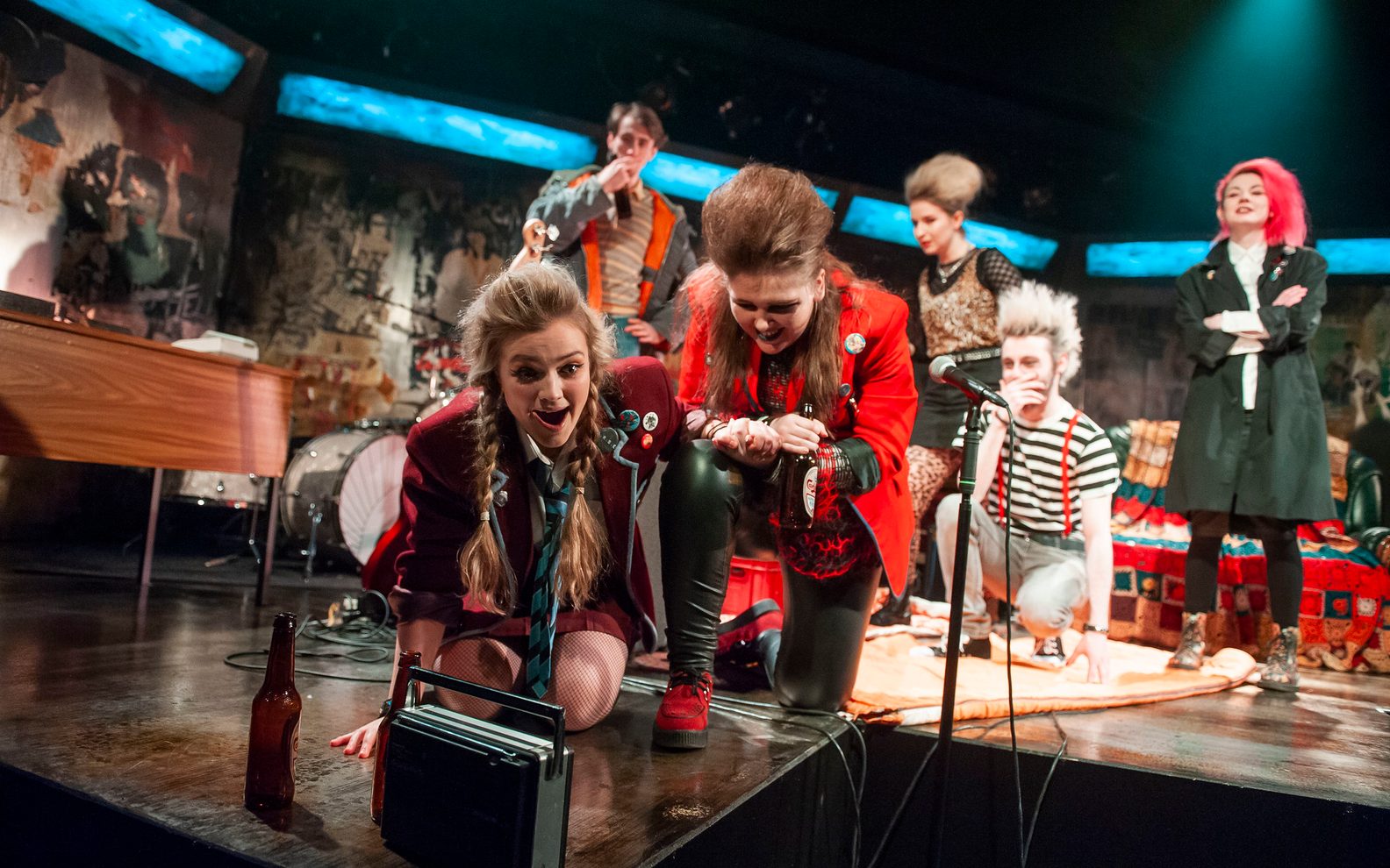 A highly theatrical production image of a show. Everybody is wearing punk style clothing. They are on a stage with graffiti style surroundings. A group of young people are crouched round an 70s style radio.