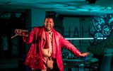 A highly theatrical production Image of a theatre show. A man wearing a red blazer, brown trousers and a red scarf is smiling and dancing.