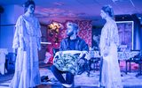 A highly theatrical production Image of a theatre show. One actor is sitting on a chair tied up whilst two women stand either side of him looking at each other.