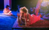 A highly theatrical production Image of a theatre show. A girl in old, dirty, victoriam clothes is lying on the floor holding the temples of her head.