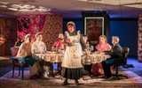 A highly theatrical production Image of a theatre show. A group of actors in old, dishevelled, Victorian style clothing are sitting round a large dining table.